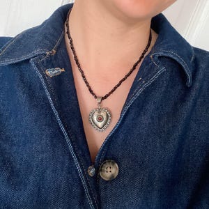 May include: A silver heart pendant necklace with a small red stone at its centre. The pendant is suspended from a beaded necklace. The person is wearing a dark blue denim jacket.