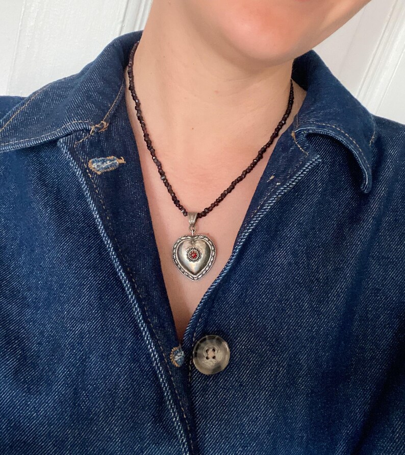 May include: A silver heart pendant necklace with a small red stone at its centre. The pendant is suspended from a beaded necklace. The person is wearing a dark blue denim jacket.