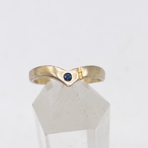 May include: A gold V-shaped ring featuring a single small round blue gemstone set at the centre point. The band has a smooth, matte finish and includes a small vertical etched line detail on the right side of the chevron peak.