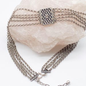 May include: A silver-toned choker necklace with multiple strands of delicate chain. The necklace features a rectangular centerpiece with an intricate, openwork design. The clasp and adjustable chain complete the piece, showcasing a vintage aesthetic.