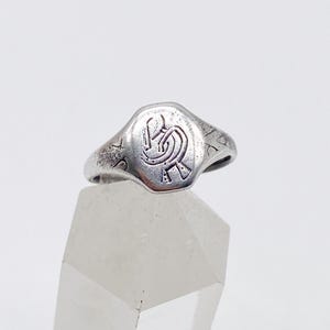 May include: A silver signet ring with a detailed, raised design on the oval face. The ring is set against a white background, showcasing its vintage style. The ring's design appears to be a stylized monogram or emblem.
