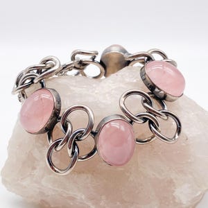 Puede incluir: Una pulsera de plata con piedras preciosas ovaladas de color rosa p&aacute;lido. Las piedras est&aacute;n engastadas en biseles de plata y unidas por eslabones de cadena de plata. La pulsera se muestra sobre una superficie rosa claro texturizada.