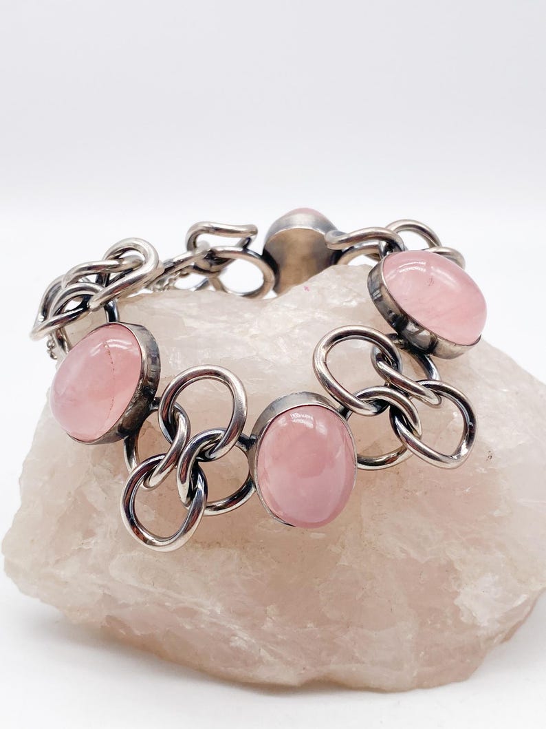 Puede incluir: Una pulsera de plata con piedras preciosas ovaladas de color rosa p&aacute;lido. Las piedras est&aacute;n engastadas en biseles de plata y unidas por eslabones de cadena de plata. La pulsera se muestra sobre una superficie rosa claro texturizada.