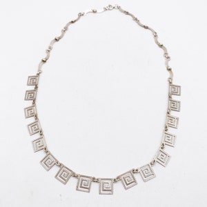 May include: A silver necklace with a chain made of small, wavy links. The necklace has a series of square pendants with a geometric design.