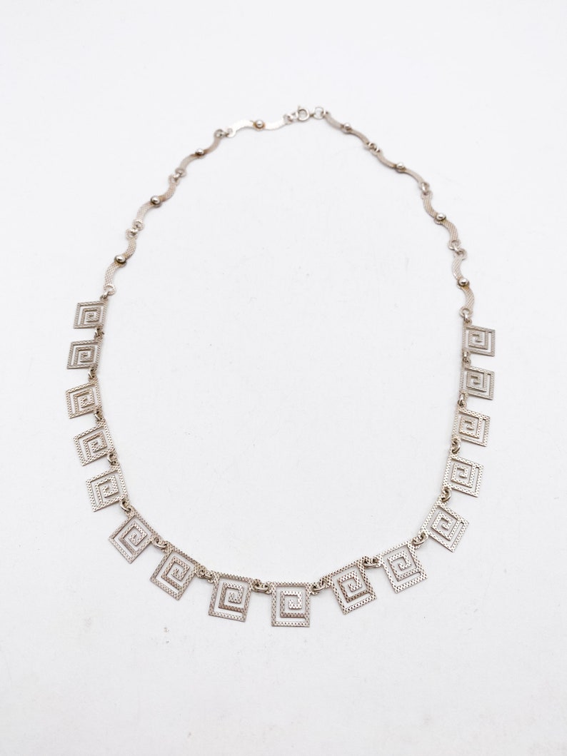 May include: A silver necklace with a chain made of small, wavy links. The necklace has a series of square pendants with a geometric design.