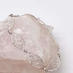 May include: A silver bracelet with a delicate, intricate design. The bracelet is made up of a series of oval-shaped links, each with a geometric pattern.