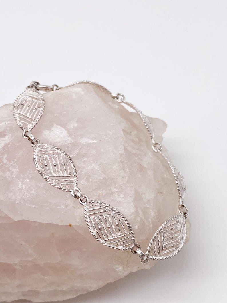May include: A silver bracelet with a delicate, intricate design. The bracelet is made up of a series of oval-shaped links, each with a geometric pattern.