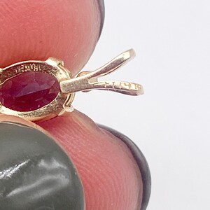 May include: Close-up of a gold pendant with an oval-shaped, deep red gemstone. The pendant has a bail for a chain and is held by a person's fingers. The metal is stamped with markings.