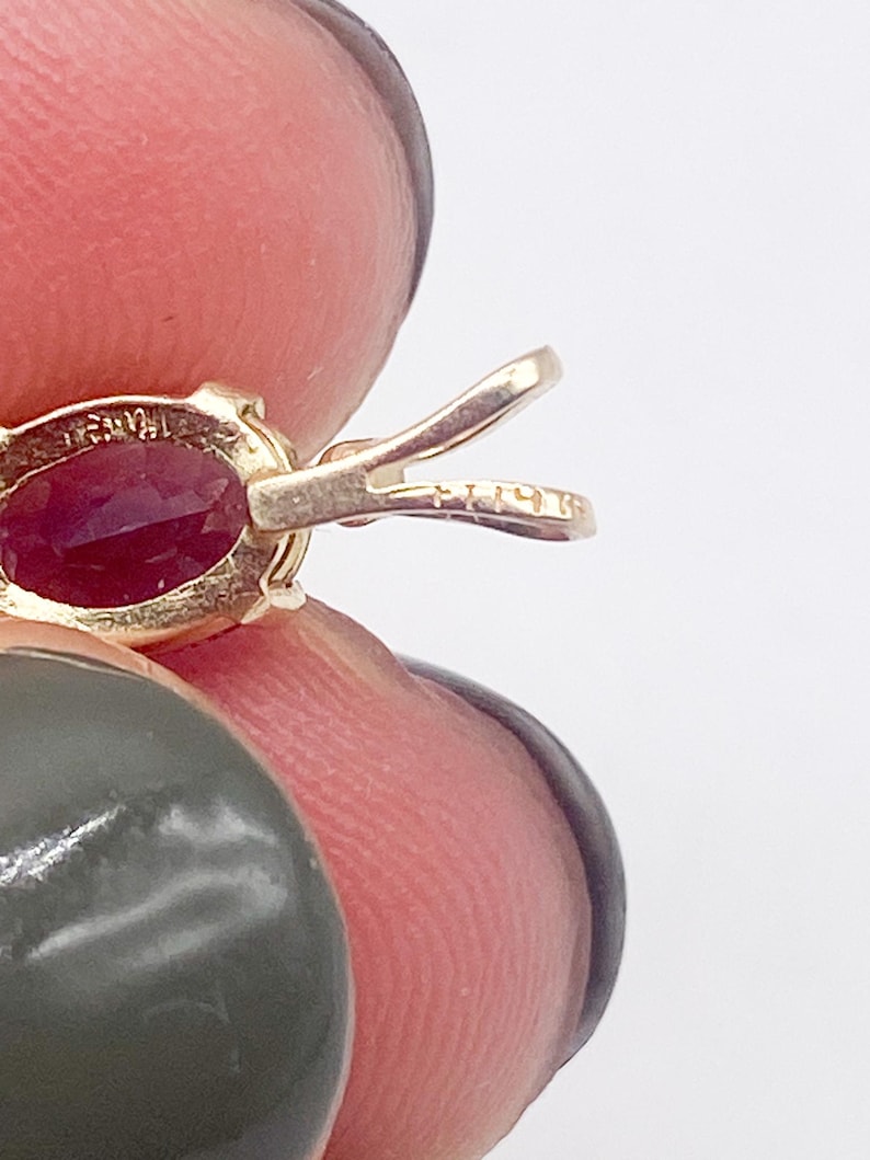 May include: Close-up of a gold pendant with an oval-shaped, deep red gemstone. The pendant has a bail for a chain and is held by a person's fingers. The metal is stamped with markings.
