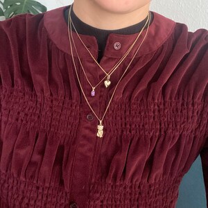 May include: Three gold necklaces with different pendants. One necklace has a small purple gemstone, another has a heart-shaped pendant, and the third has a bear pendant. The necklaces are layered and worn over a burgundy corduroy shirt.