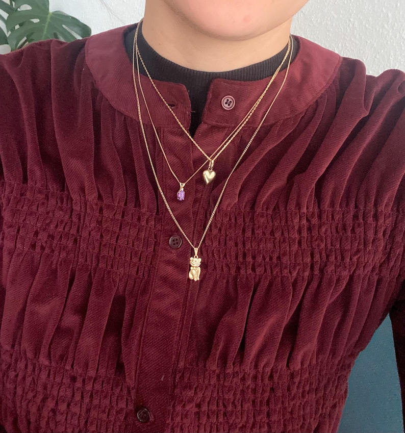 May include: Three gold necklaces with different pendants. One necklace has a small purple gemstone, another has a heart-shaped pendant, and the third has a bear pendant. The necklaces are layered and worn over a burgundy corduroy shirt.