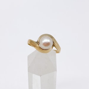 May include: A gold ring featuring a large, round, off-white pearl. The ring's band has a flowing, wave-like design that cradles the pearl. The ring is displayed on a clear, geometric stand against a white background.
