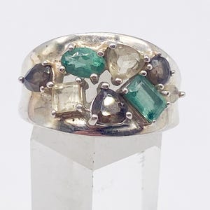 May include: A silver ring featuring an array of colorful gemstones. The ring's design includes emerald green, clear, and smoky-colored stones in various shapes. The ring is displayed on a clear, geometric stand.