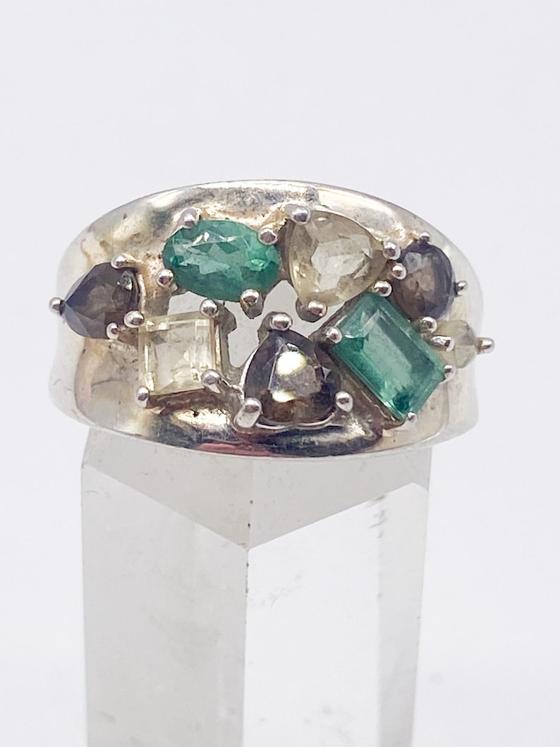 May include: A silver ring featuring an array of colorful gemstones. The ring's design includes emerald green, clear, and smoky-colored stones in various shapes. The ring is displayed on a clear, geometric stand.