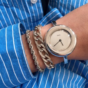 May include: A silver cuff bracelet watch with a white face and the word "Obey" printed on it. The watch is worn on a wrist with two silver bracelets.
