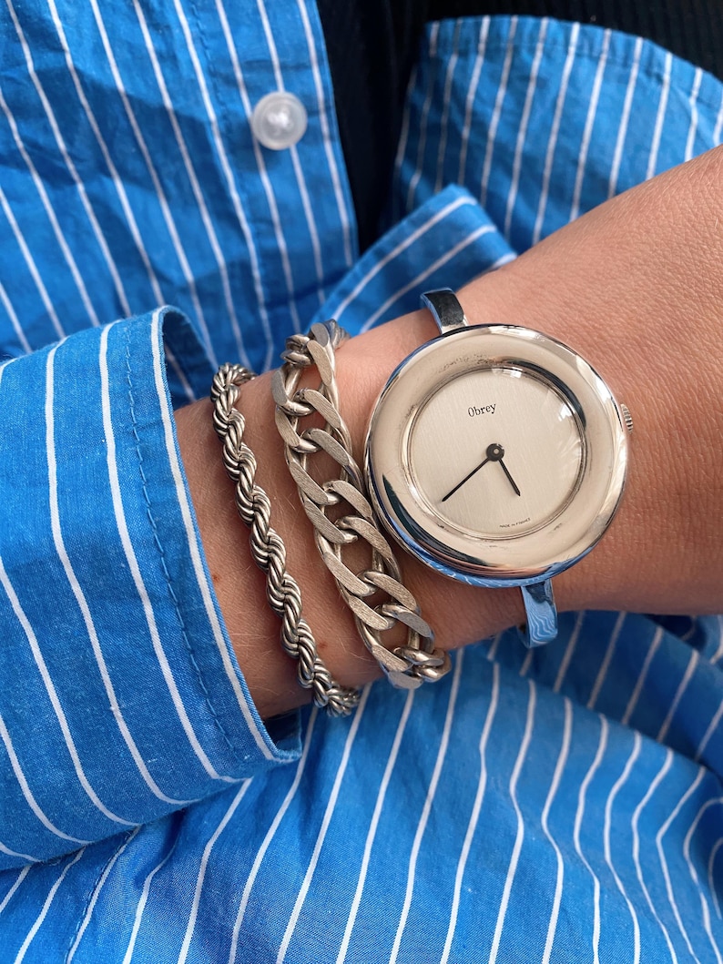 May include: A silver cuff bracelet watch with a white face and the word "Obey" printed on it. The watch is worn on a wrist with two silver bracelets.