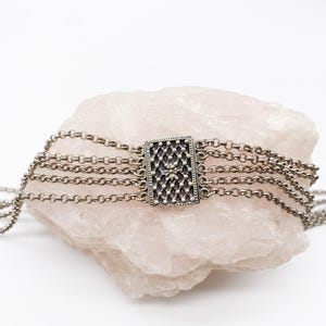 May include: A silver-toned choker necklace with multiple chain strands. The focal point is a rectangular centerpiece with a detailed, dark pattern. The necklace is displayed on a light pink, textured stone against a white background.