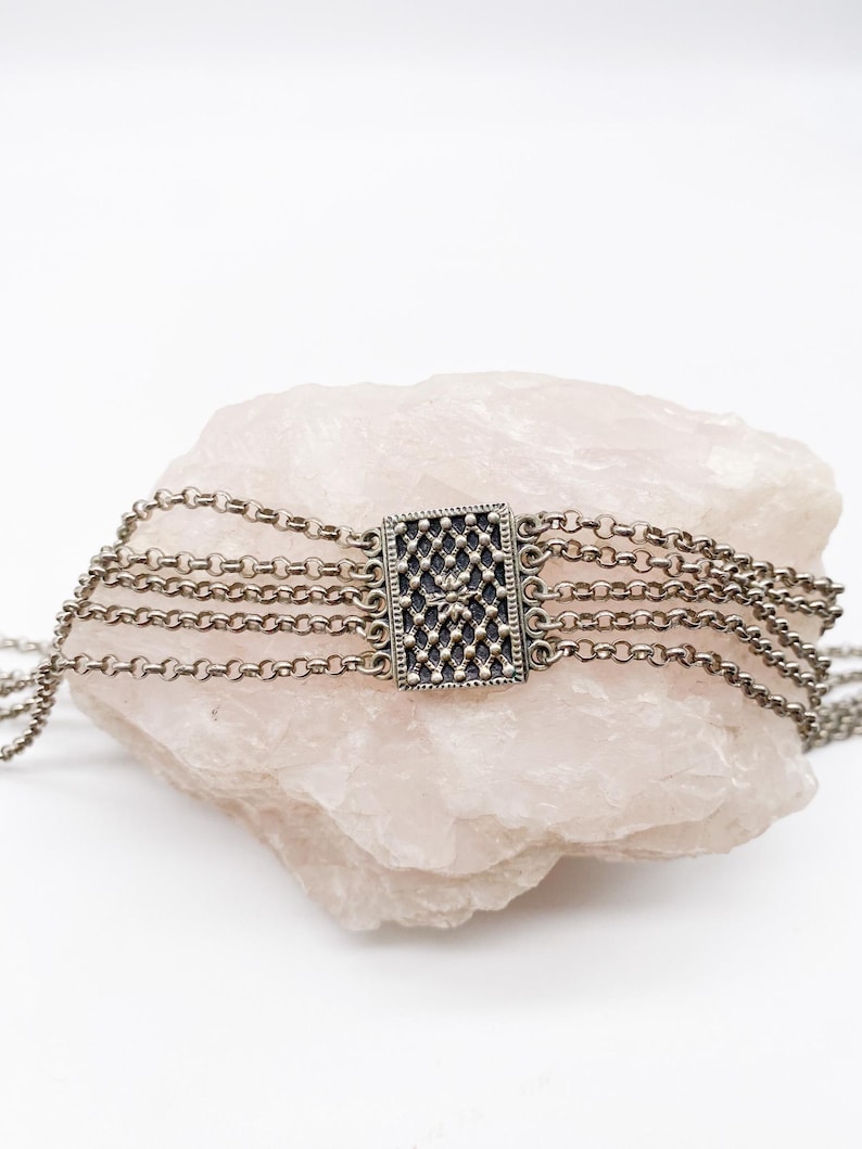 May include: A silver-toned choker necklace with multiple chain strands. The focal point is a rectangular centerpiece with a detailed, dark pattern. The necklace is displayed on a light pink, textured stone against a white background.