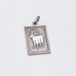 May include: A silver-toned rectangular pendant featuring a detailed ram illustration. The ram is centered on a textured background within a rectangular frame. The pendant has a small loop at the top for a chain.