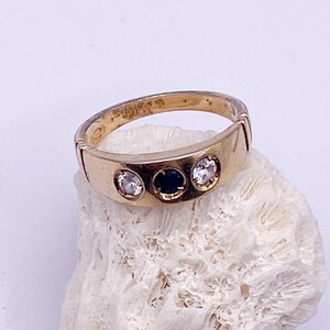 May include: A gold ring featuring three stones: two clear and one dark blue. The ring is set on a white, textured surface. The ring's band has a simple, classic design.