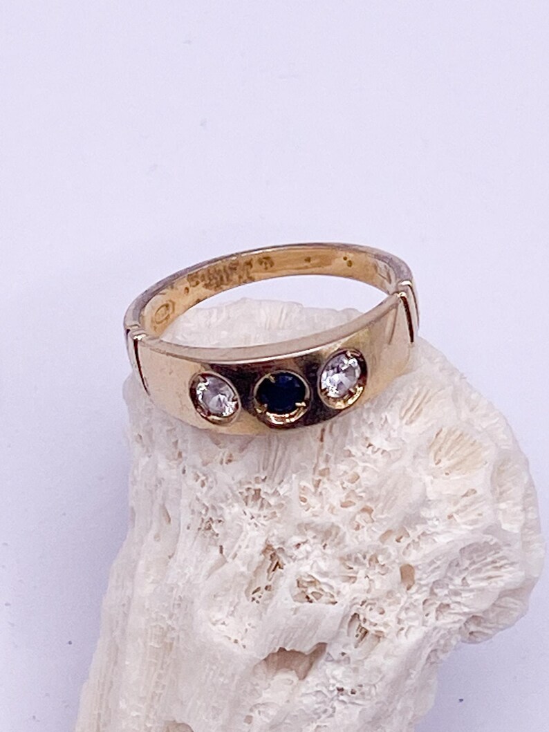 May include: A gold ring featuring three stones: two clear and one dark blue. The ring is set on a white, textured surface. The ring's band has a simple, classic design.