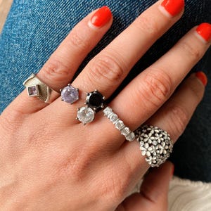 May include: A hand displaying various rings. These include a silver ring with a square purple stone, a light purple gemstone ring, a black gemstone ring, a ring with multiple clear stones, and a floral ring.