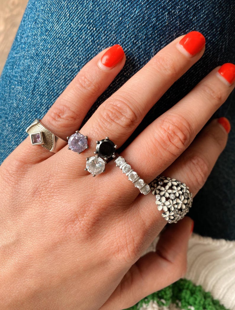 May include: A hand displaying various rings. These include a silver ring with a square purple stone, a light purple gemstone ring, a black gemstone ring, a ring with multiple clear stones, and a floral ring.