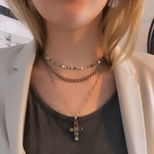 K&ouml;nnte beinhalten: Eine mehrlagige Halskette mit einer goldenen Kette und einem Kreuzanh&auml;nger mit bunten Steinen. Der Kreuzanh&auml;nger befindet sich in der Mitte des Bildes. Die Halskette wird &uuml;ber einem grauen T-Shirt getragen.