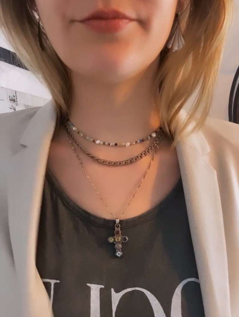 K&ouml;nnte beinhalten: Eine mehrlagige Halskette mit einer goldenen Kette und einem Kreuzanh&auml;nger mit bunten Steinen. Der Kreuzanh&auml;nger befindet sich in der Mitte des Bildes. Die Halskette wird &uuml;ber einem grauen T-Shirt getragen.