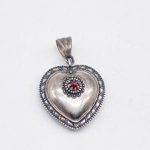 May include: A silver heart-shaped pendant with a small red gemstone at its centre. The pendant features a detailed border and a loop for a chain. The heart is slightly domed, giving it a three-dimensional appearance.