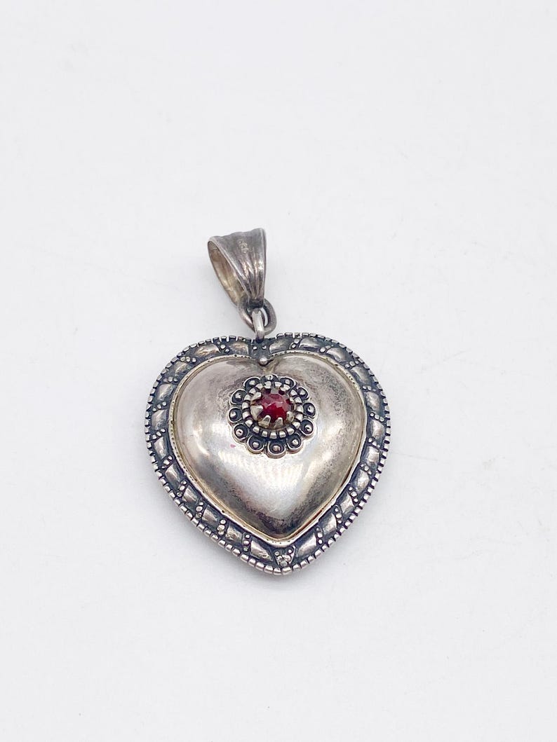 May include: A silver heart-shaped pendant with a small red gemstone at its centre. The pendant features a detailed border and a loop for a chain. The heart is slightly domed, giving it a three-dimensional appearance.
