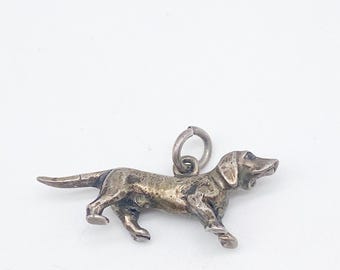May include: A small, silver-toned dachshund charm. The dog is depicted in a walking pose, with detailed features. The charm has a loop at the top for attachment. The charm is approximately 2.5 cm long.