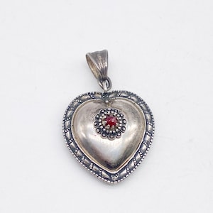 May include: A silver heart pendant with a central red gemstone. The pendant features a detailed border and a loop for a chain. The heart shape is slightly domed, creating a three-dimensional effect.