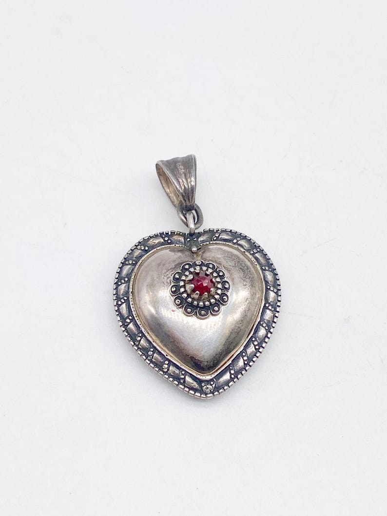 May include: A silver heart pendant with a central red gemstone. The pendant features a detailed border and a loop for a chain. The heart shape is slightly domed, creating a three-dimensional effect.