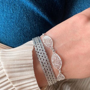 May include: Two silver bracelets on a wrist. One bracelet is a delicate chain with oval-shaped links that have a geometric pattern. The other bracelet is a thicker, more substantial chain with a square-shaped pattern.