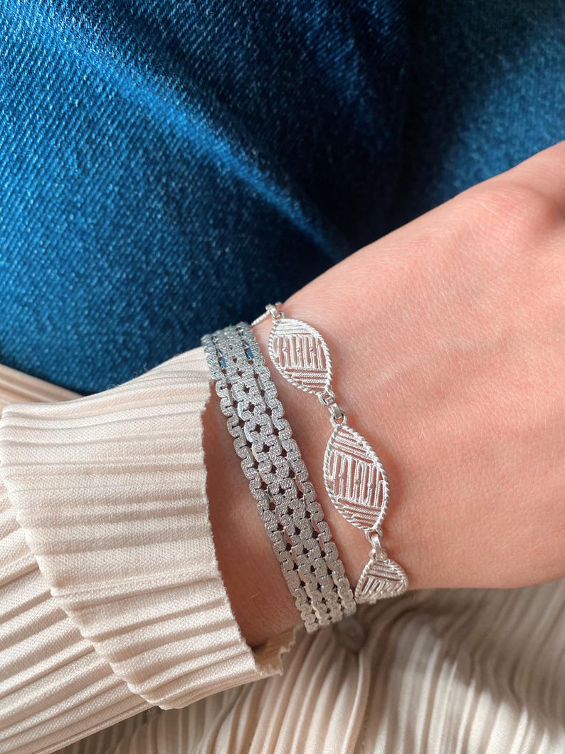 May include: Two silver bracelets on a wrist. One bracelet is a delicate chain with oval-shaped links that have a geometric pattern. The other bracelet is a thicker, more substantial chain with a square-shaped pattern.