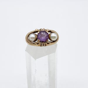 May include: A gold ring featuring a central oval amethyst gemstone flanked by two round pearls. The ring has an ornate design with delicate detailing around the gemstones. The ring is displayed on a clear, faceted stand.