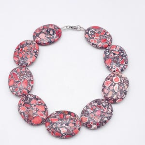 May include: A necklace composed of nine large, oval-shaped beads featuring a mottled pattern of deep red, pink, and dark gray fragments. The beads are linked together in a circular arrangement and secured with a small silver-toned metal clasp.