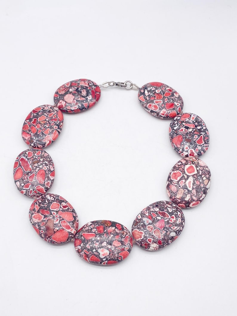 May include: A necklace composed of nine large, oval-shaped beads featuring a mottled pattern of deep red, pink, and dark gray fragments. The beads are linked together in a circular arrangement and secured with a small silver-toned metal clasp.