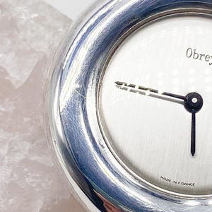 May include: A silver pocket watch with a white face and black hands. The watch face has the text "Obrey" and "Made in France".