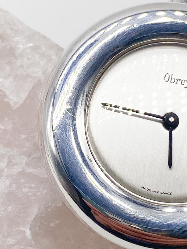 May include: A silver pocket watch with a white face and black hands. The watch face has the text "Obrey" and "Made in France".
