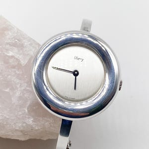 May include: A silver Obry wristwatch with a round face and a silver bangle-style band. The watch face has a white background with black hands and the word "Obry" printed in black.