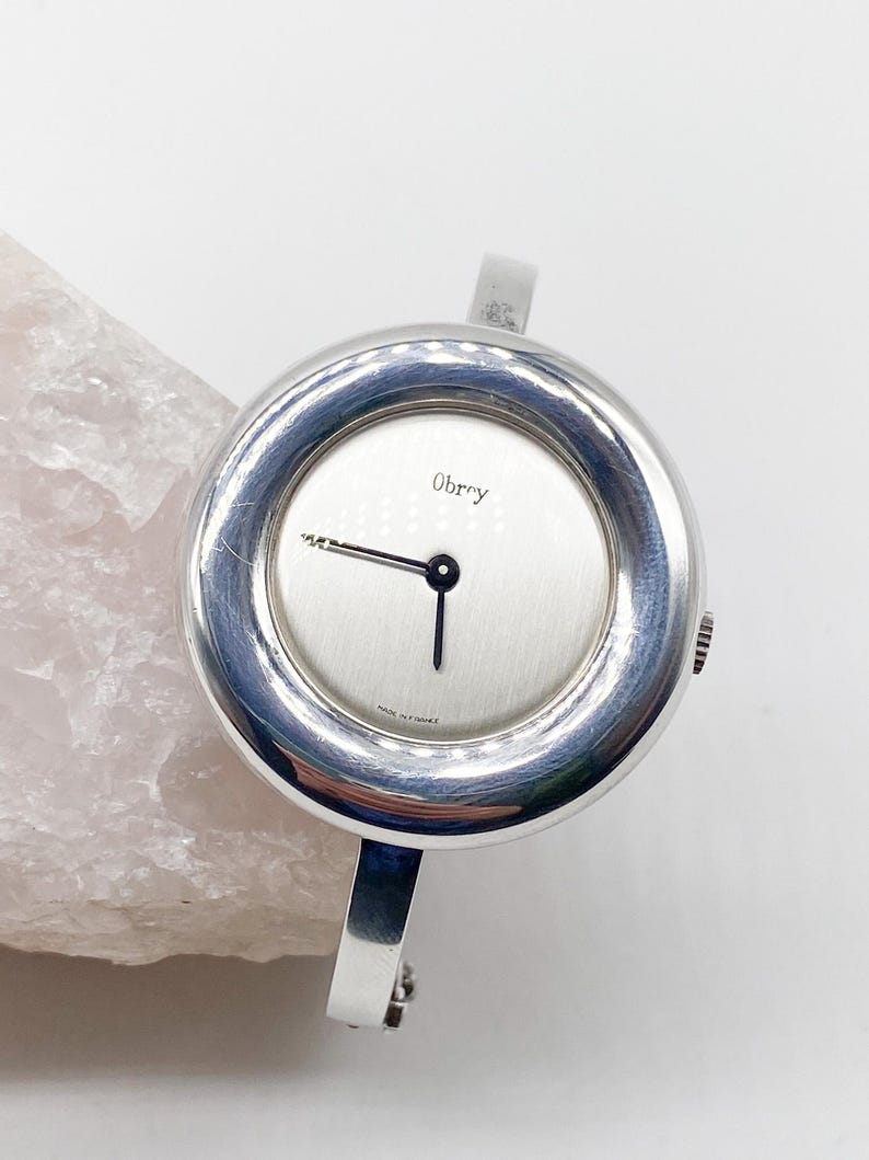 May include: A silver Obry wristwatch with a round face and a silver bangle-style band. The watch face has a white background with black hands and the word "Obry" printed in black.