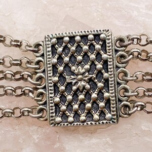 May include: Close-up of a silver-toned bracelet featuring a rectangular centerpiece with a detailed, openwork design. The bracelet has multiple strands of delicate chain links, creating a layered effect. The centerpiece has a floral design.