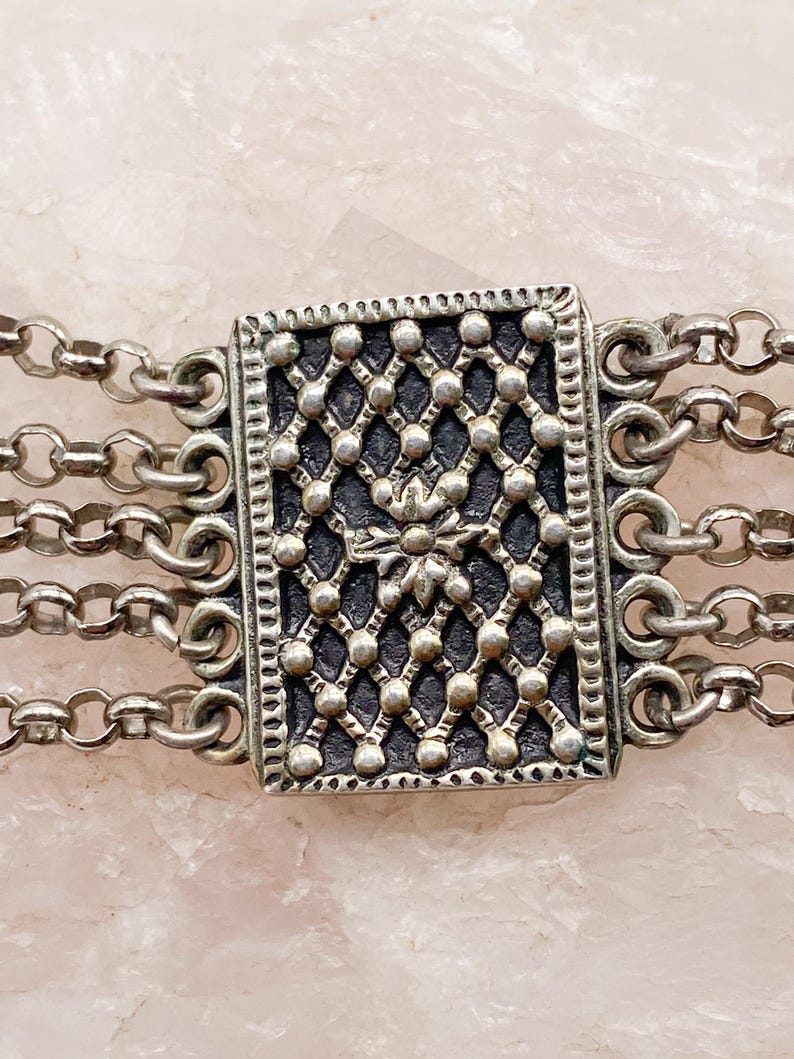 May include: Close-up of a silver-toned bracelet featuring a rectangular centerpiece with a detailed, openwork design. The bracelet has multiple strands of delicate chain links, creating a layered effect. The centerpiece has a floral design.