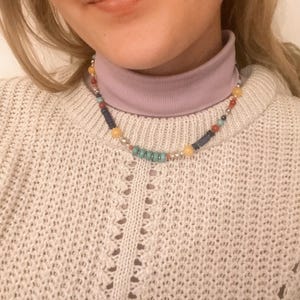 Pu&ograve; includere: Una collana di perline multicolori con perline gialle, blu, turchesi e rosse. La collana &egrave; indossata da una persona con un maglione a collo alto viola chiaro e un maglione di lana bianco.