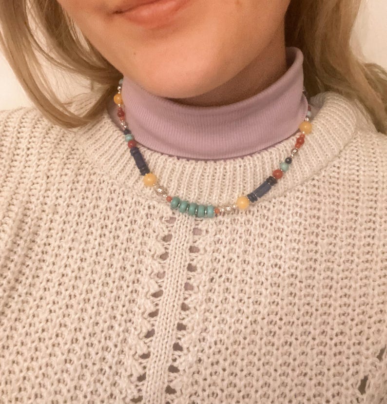 Pu&ograve; includere: Una collana di perline multicolori con perline gialle, blu, turchesi e rosse. La collana &egrave; indossata da una persona con un maglione a collo alto viola chiaro e un maglione di lana bianco.