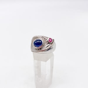 May include: A silver ring featuring a large, oval, deep blue cabochon stone and a smaller, round, vibrant pink gemstone. The metal band has a textured, ridged surface detail on one side, creating a contrast with the smooth, polished finish of the silver.