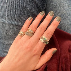 May include: A hand with olive green nail polish, adorned with four gold rings. One ring features a row of small diamonds, another has a cluster of green stones, and the others have unique designs. The rings are set against a backdrop of denim and burgundy fabric.