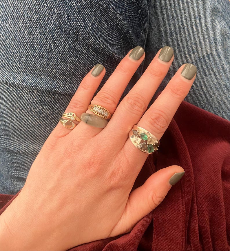 May include: A hand with olive green nail polish, adorned with four gold rings. One ring features a row of small diamonds, another has a cluster of green stones, and the others have unique designs. The rings are set against a backdrop of denim and burgundy fabric.